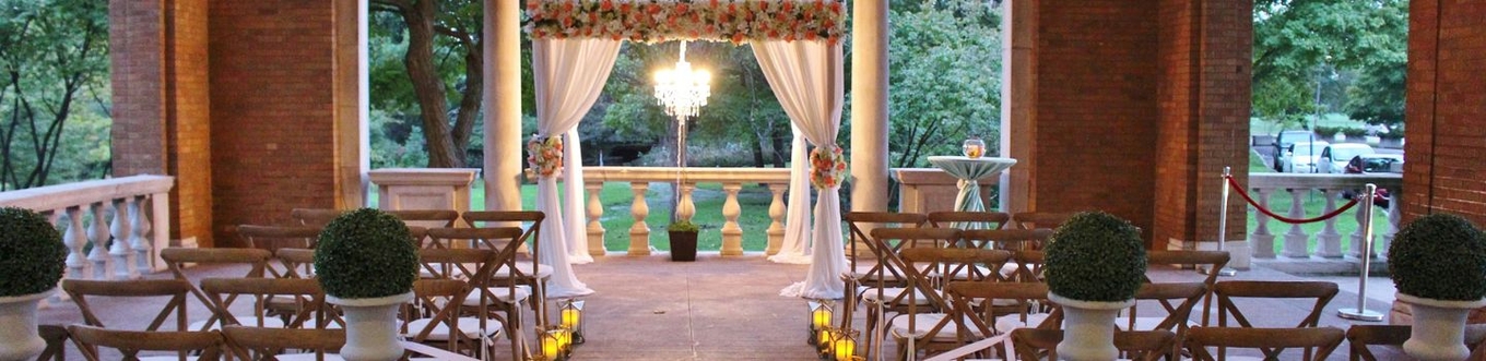 Outdoor wedding venue decorated with floral arrangements, white draping, and wooden cross-back chairs.